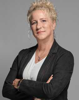 A confident woman with curly hair stands with arms crossed, wearing a black jacket and white shirt against a gray background.