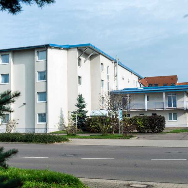 A modern residential building with blue accents stands surrounded by greenery on a quiet street.