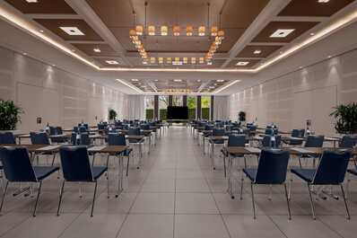 A spacious meeting room with rows of tables and chairs is set up for a conference, featuring a large screen at the front.