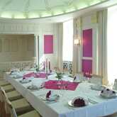 An elegant dining room with a long table set for a formal event, featuring pink accents and decorative columns.