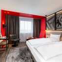 Modern hotel room with red accents features a double bed, a chair, a TV, and a stylish desk with a kettle and water bottle.