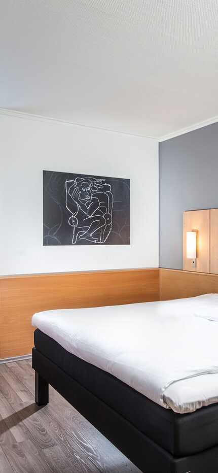 A modern single room features a cozy bed, a bright red chair by the window, and a stylish wall art piece.