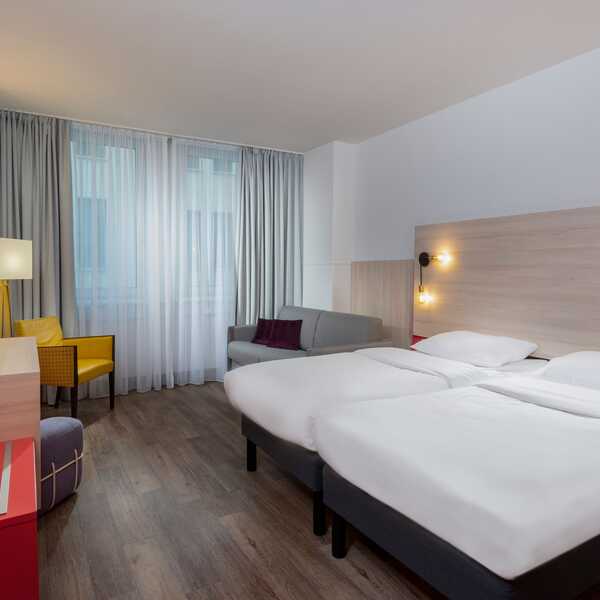Helles Hotelzimmer mit zwei Betten, einem grauen Sofa und gelbem Stuhl bietet Komfort für Familienaufenthalte.