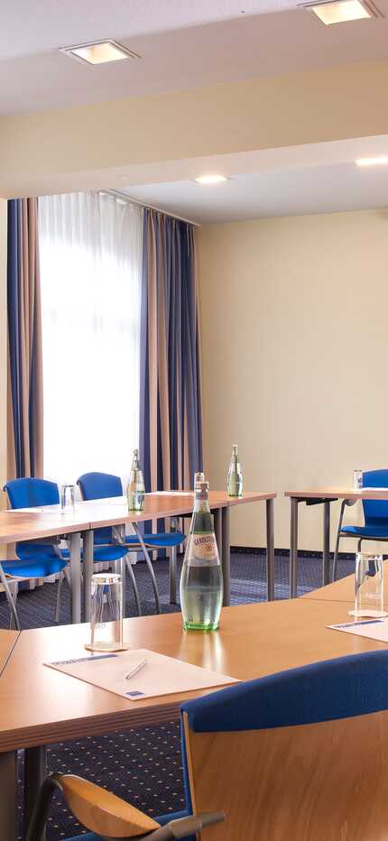 Ein heller Konferenzraum mit U-förmiger Tischanordnung, blau gepolsterten Stühlen und bereitgestellten Wasserflaschen.