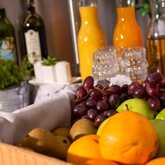 A breakfast spread features a wooden crate of fresh grapes, oranges, and apples, with juice bottles and herbs in the background.