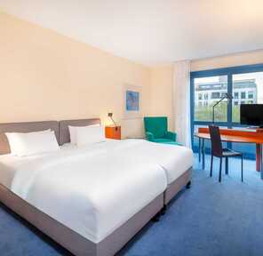 A modern hotel room features a cozy double bed, a desk with a TV, and a large window offering city views.