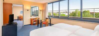 A bright hotel room features twin beds, a TV, and a dining area, with large windows offering a scenic city view.