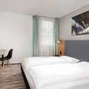 A modern double room features a neatly made bed, a desk with a chair, and an industrial artwork on the wall.