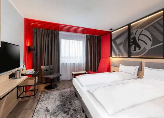 A modern hotel room features a double bed, stylish decor, and a seating area by a window with red accents.