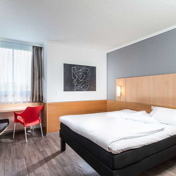 Ein modernes Einzelzimmer mit einem großen Bett, einem roten Stuhl am Schreibtisch und einem Kunstwerk an der Wand.