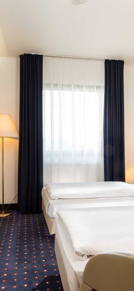 A bright twin bed room features modern decor, a wall-mounted TV, and ample natural light from a large window.
