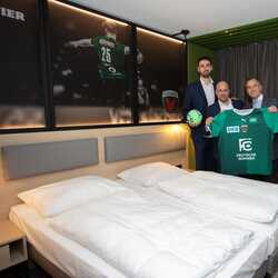 Four men in suits hold a green sports jersey and a ball in a hotel room, showcasing team spirit.