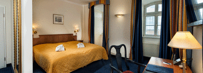 A cozy classic room features a warm yellow bedspread, elegant striped curtains, and a wooden desk with a lamp.