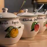 Eine Reihe von vier weißen, keramischen Marmeladentöpfen mit Deckeln und kleinen Löffeln. Jeder Topf ist handbemalt mit einer Frucht und dem Namen des Fruchtaufstrichs auf Deutsch, wie "Marille - Apricot" und "Weichsel - Morello Cherry".