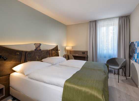 A cozy double room features a stylish headboard, a neatly made bed, a desk with a lamp, and a window with light curtains.