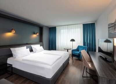 A modern hotel room with a double bed, blue armchair, and desk, illuminated by stylish pendant lights.