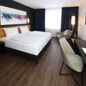 A modern hotel room features a sleek double bed, abstract art, and a workspace with a smooth desk and chair.