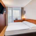 A cozy hotel room features a double bed, wall-mounted TV, and a window with sheer curtains, creating a relaxing ambiance.