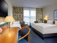Ein gemütliches Hotelzimmer mit Doppelbett, Schreibtisch, Fernseher und blumigen Vorhängen strahlt Komfort aus.