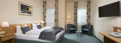 A modern double room features a cozy bed, two armchairs, a flat-screen TV, and decorative curtains, creating a welcoming atmosphere.