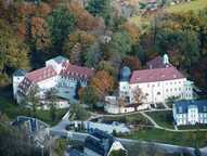 Luftaufnahme eines großen Gebäudekomplexes umgeben von herbstlich verfärbtem Wald in einer idyllischen Landschaft.