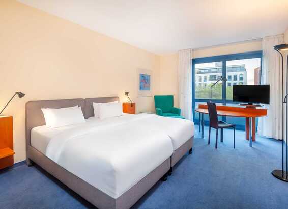 A modern hotel room features a cozy double bed, a desk with a TV, and a large window offering city views.