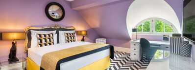 A stylish boutique room features a plush bed, purple walls, and a chevron-patterned floor under a round window.