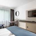 A modern hotel room features a sleek TV, a cozy chair, and a desk by a large window with sheer curtains.