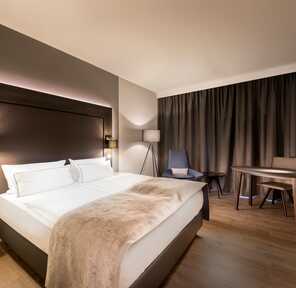 A modern hotel room features a cozy bed, sleek furniture, and dark curtains, creating an inviting and relaxing atmosphere.
