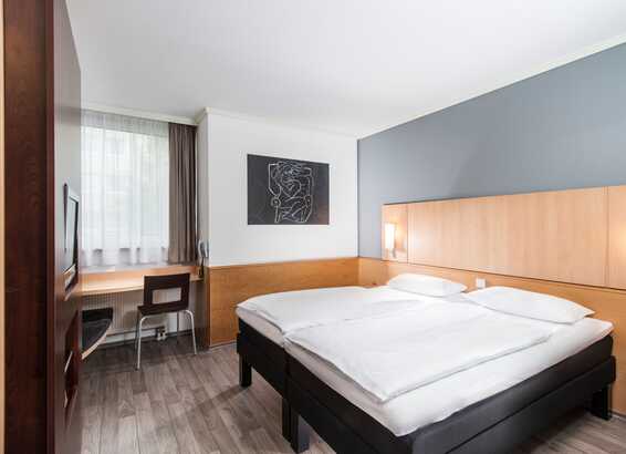A modern double room features a neatly made bed, artwork, and a desk by a window, creating a cozy and inviting atmosphere.