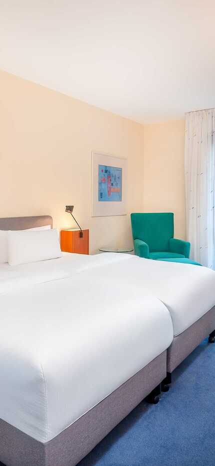 A modern hotel room features a cozy double bed, a desk with a TV, and a large window offering city views.