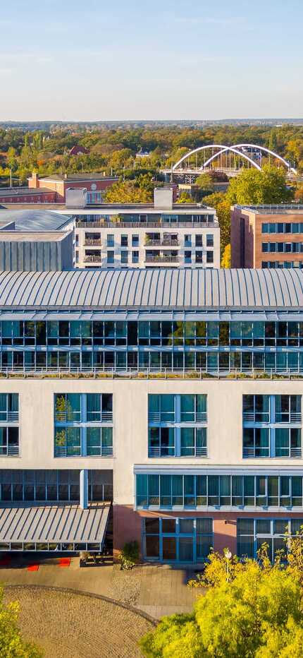 A modern, multi-story building with large windows stands amidst a scenic urban landscape under a clear blue sky.