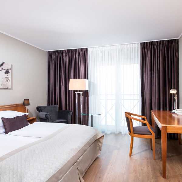 Ein modernes Standard-Doppelzimmer mit Doppelbett, Schreibtisch, Sessel und großen Fenstern mit Vorhängen.
