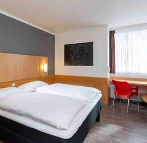 Ein modernes Standardzimmer mit einem Doppelbett, einem Schreibtisch und einem roten Stuhl unter einem großen Fenster.