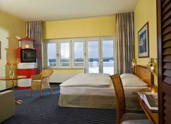 A cozy hotel room features a comfortable bed, wicker chair, and a vintage TV, with a view through large windows.