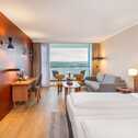 A modern hotel room features a cozy bed, a sofa, and a desk, with a scenic balcony view.