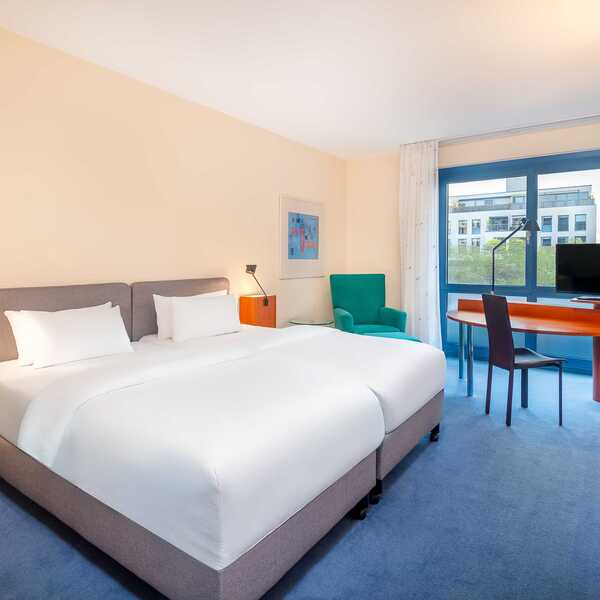 Ein modernes Hotelzimmer mit Doppelbett, Schreibtisch und Fernseher bietet einen Blick auf die Stadt durch ein großes Fenster.