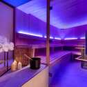 A serene sauna with glowing purple lights, wooden benches, and neatly stacked towels creates a relaxing atmosphere.