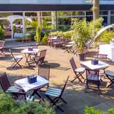 A sunlit terrace features wooden tables and chairs surrounded by lush greenery, perfect for outdoor dining.