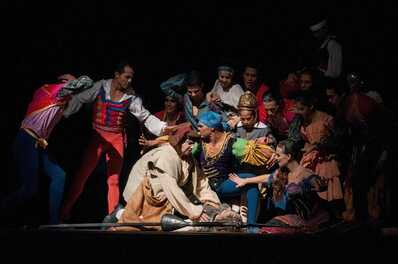 A group of performers in vibrant costumes enact a dramatic scene from "Don Quijote" on a dimly lit stage.