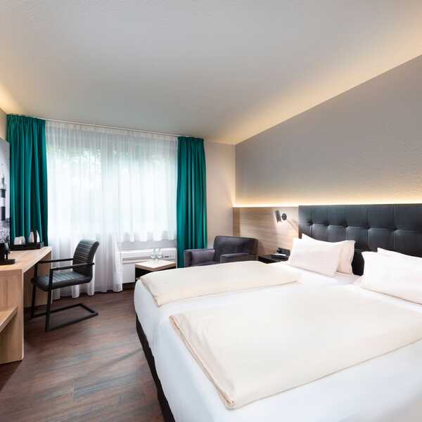 Ein modernes und komfortables Doppelzimmer im Best Western Hotel Achim Bremen. Es verfügt über zwei Einzelbetten mit dunklem Kopfteil und weißer Bettwäsche. Links vom Bett befindet sich ein Schreibtisch mit Stuhl und einem Fernseher an der Wand. Der Raum