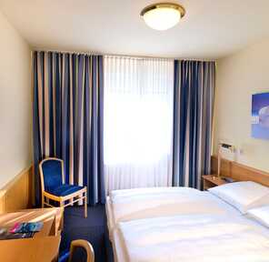 Ein Doppelzimmer mit einem gemütlichen Bett, einem Schreibtisch und blauen Vorhängen wird von Sonnenlicht durchflutet.