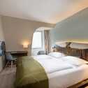 A cozy double room features a large bed with a green throw, a modern TV, and a desk with a lamp by the window.