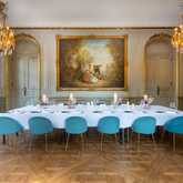 An elegant dining room features a long table with blue chairs, set under ornate chandeliers and a large classical painting.