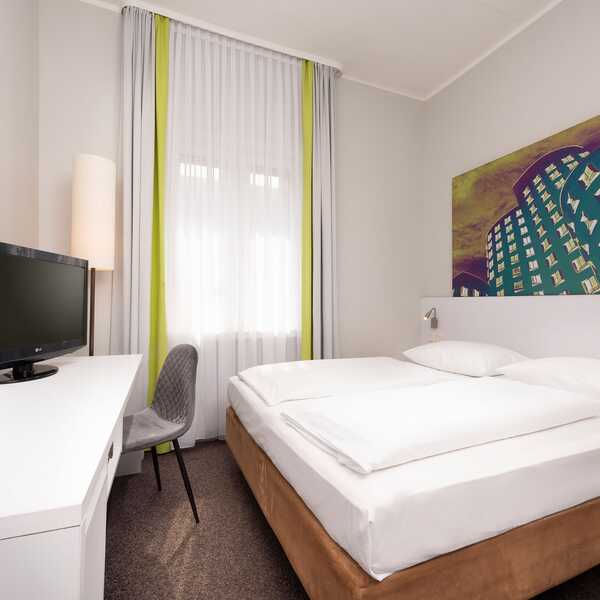 Ein modernes, helles Hotelzimmer mit Doppelbett, Flachbildfernseher und farbenfrohem Wandbild lädt zum Entspannen ein.