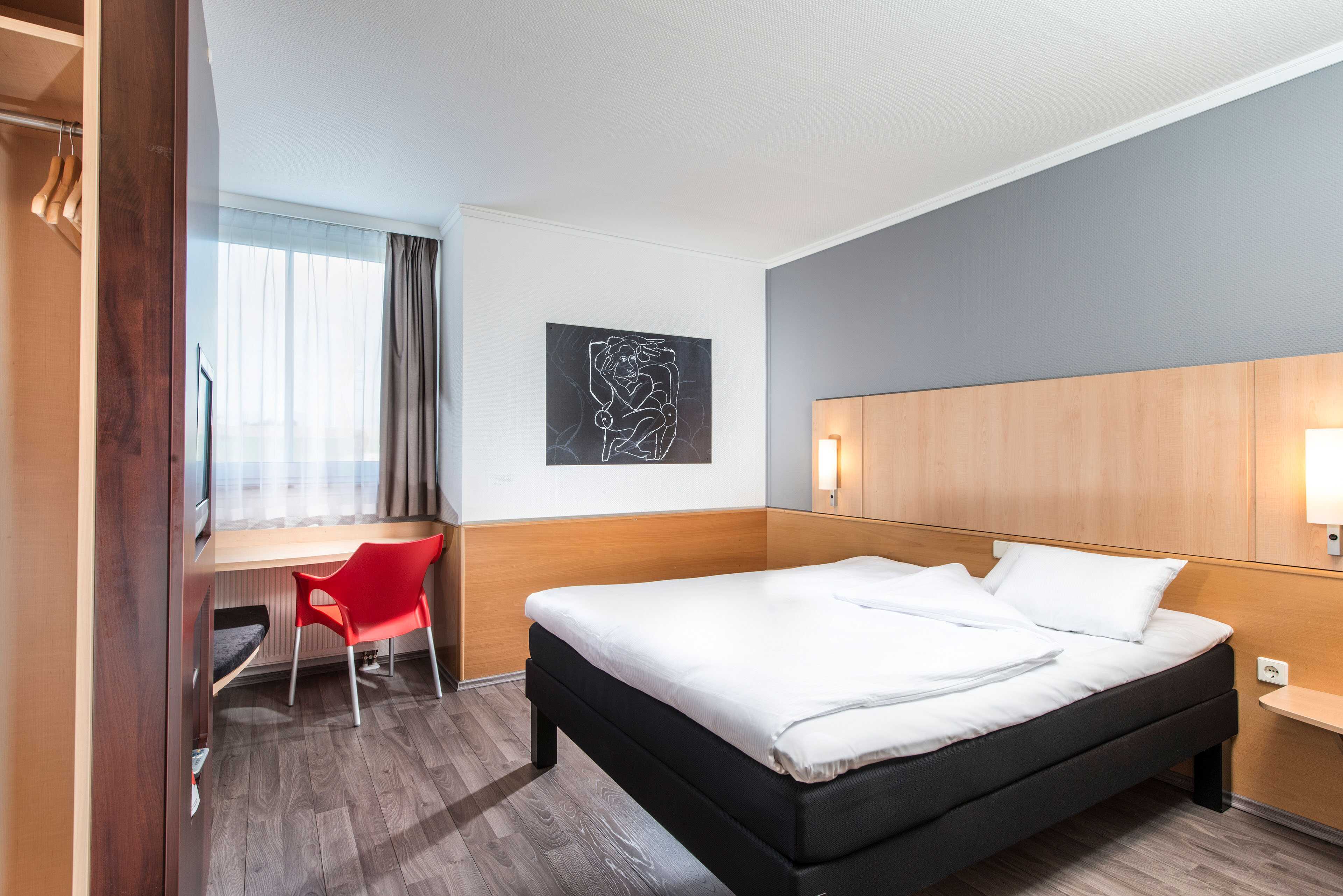 A modern single room features a cozy bed, a bright red chair by the window, and a stylish wall art piece.