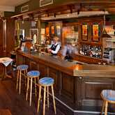 Ein Barkeeper bedient Gäste in einer gemütlichen Hotelbar mit Holzmöbeln und farbenfrohen Glasfenstern.