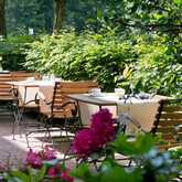 Wooden chairs and tables are set for dining on a lush, sunlit terrace surrounded by greenery and vibrant flowers.