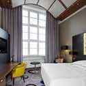 A modern hotel room features a large window, cozy bed, and stylish decor with a yellow chair and elegant lighting.