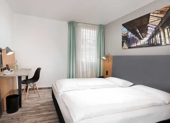 Ein modernes Doppelzimmer mit einem großen Bett, Schreibtisch und Wandkunst, das eine helle und einladende Atmosphäre bietet.